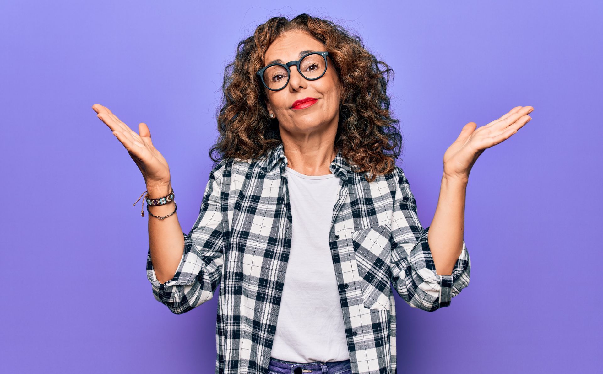 Woman with curly hair and glasses shrugs with open hands, purple background.