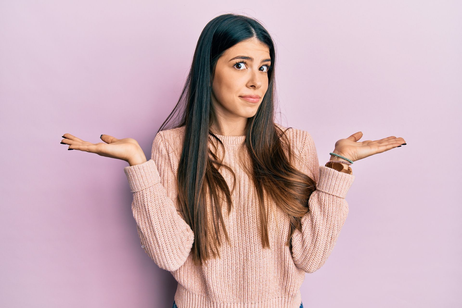 Woman with long brown hair wearing a pink sweater, shrugging with both arms outstretched and palms up.