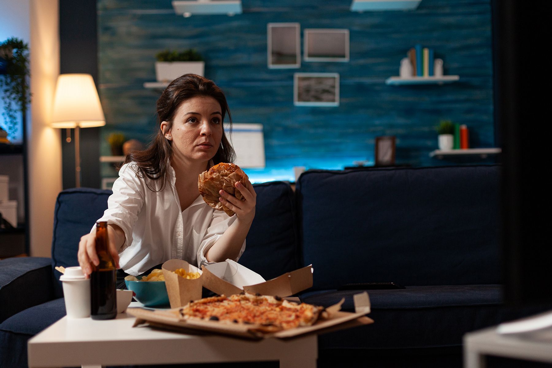 Woman eating fast food on a couch in a living room, holding a burger.