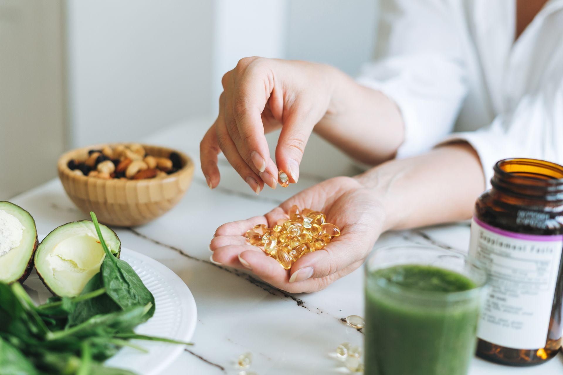 Person holding vitamins, with nuts, vegetables, and green juice on a table.