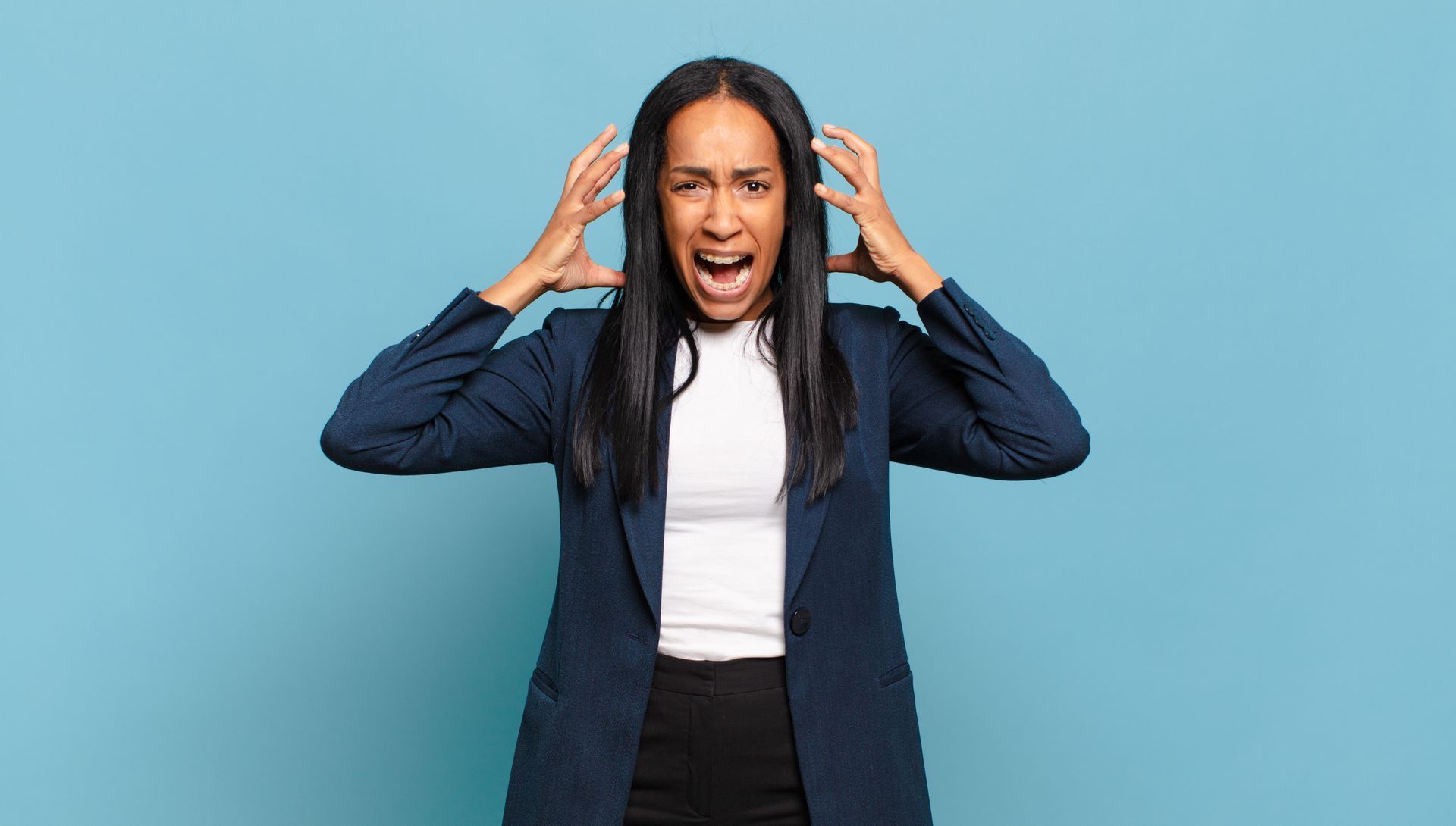 Woman in blazer, hands on head, yelling with a frustrated expression against a blue background.