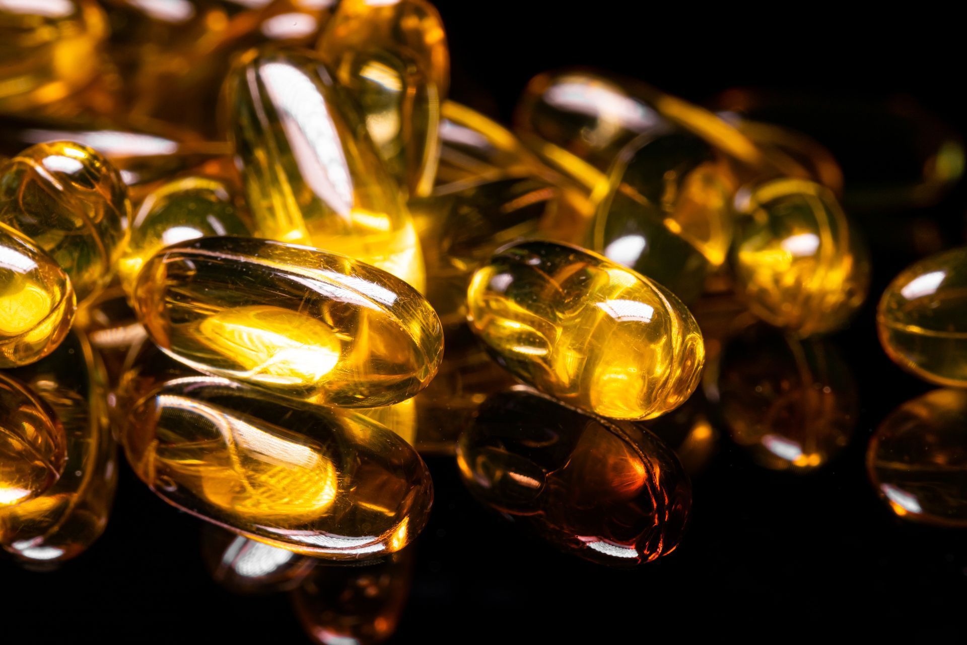 Close-up of numerous, glossy, yellow fish oil capsules on a black reflective surface.