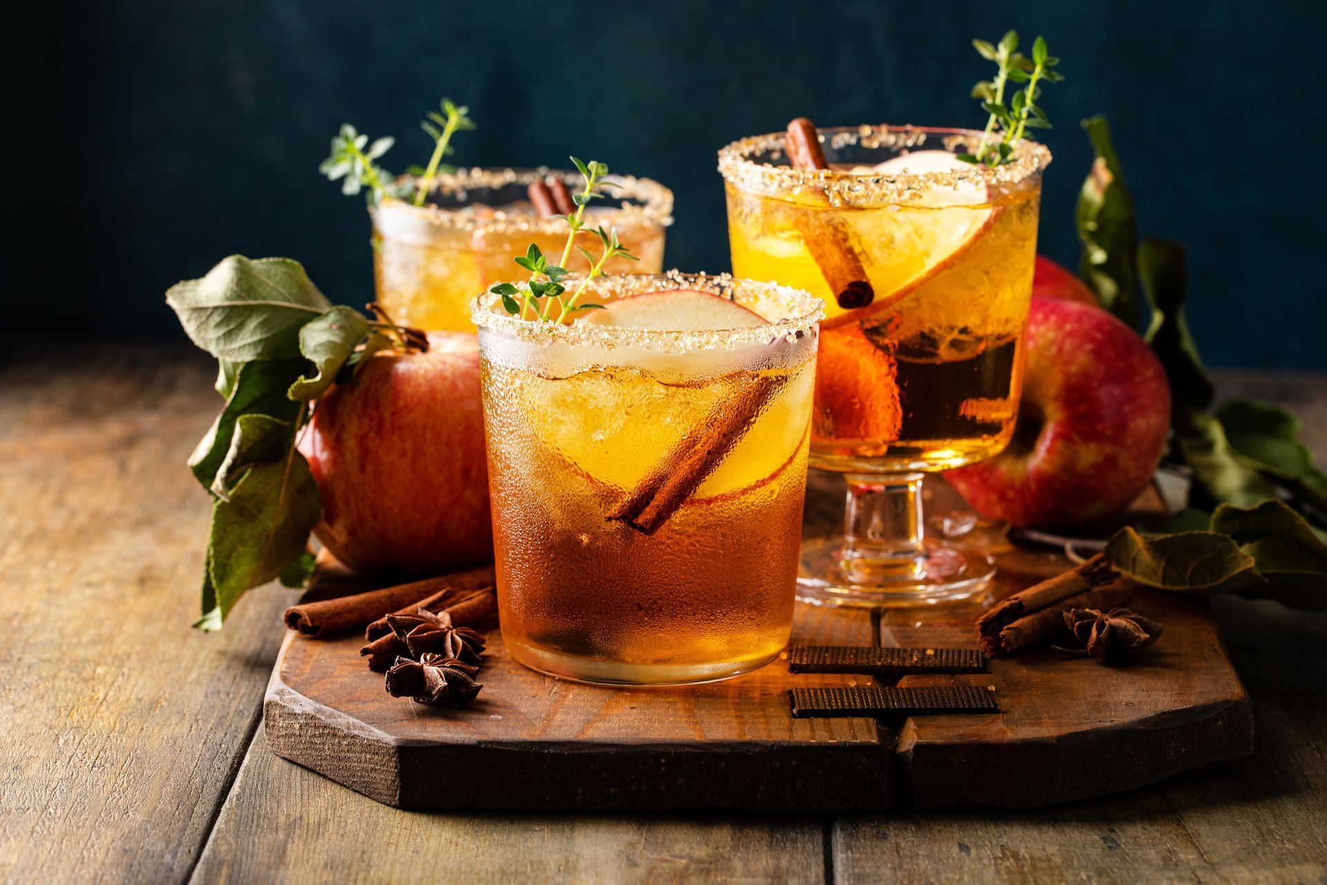 Three apple cider cocktails with cinnamon sticks and apple slices, on a wooden board with apples and spices.