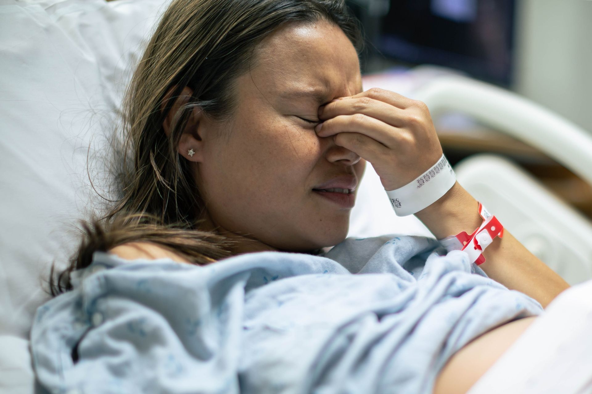 Woman in hospital bed with pained expression, hand on face. Wearing blue gown, wristband visible.