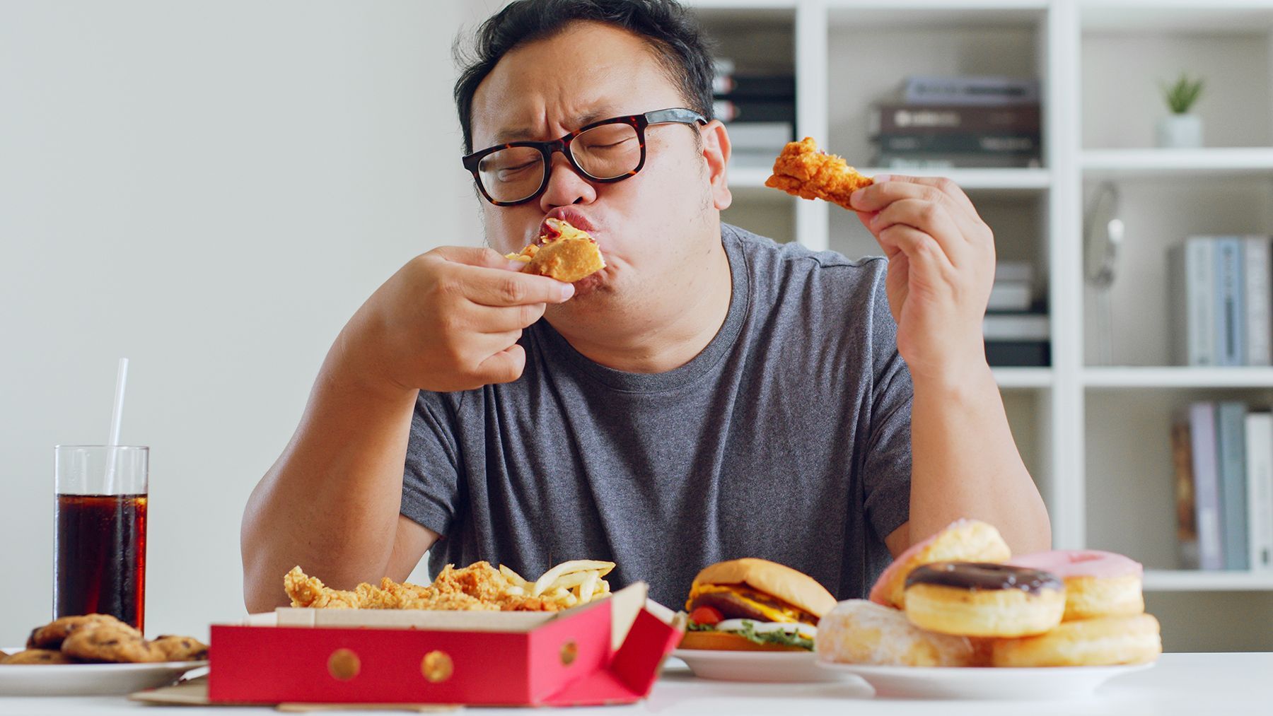 Man eating fast food, including fried chicken, burgers, donuts, and soda, at a table indoors.