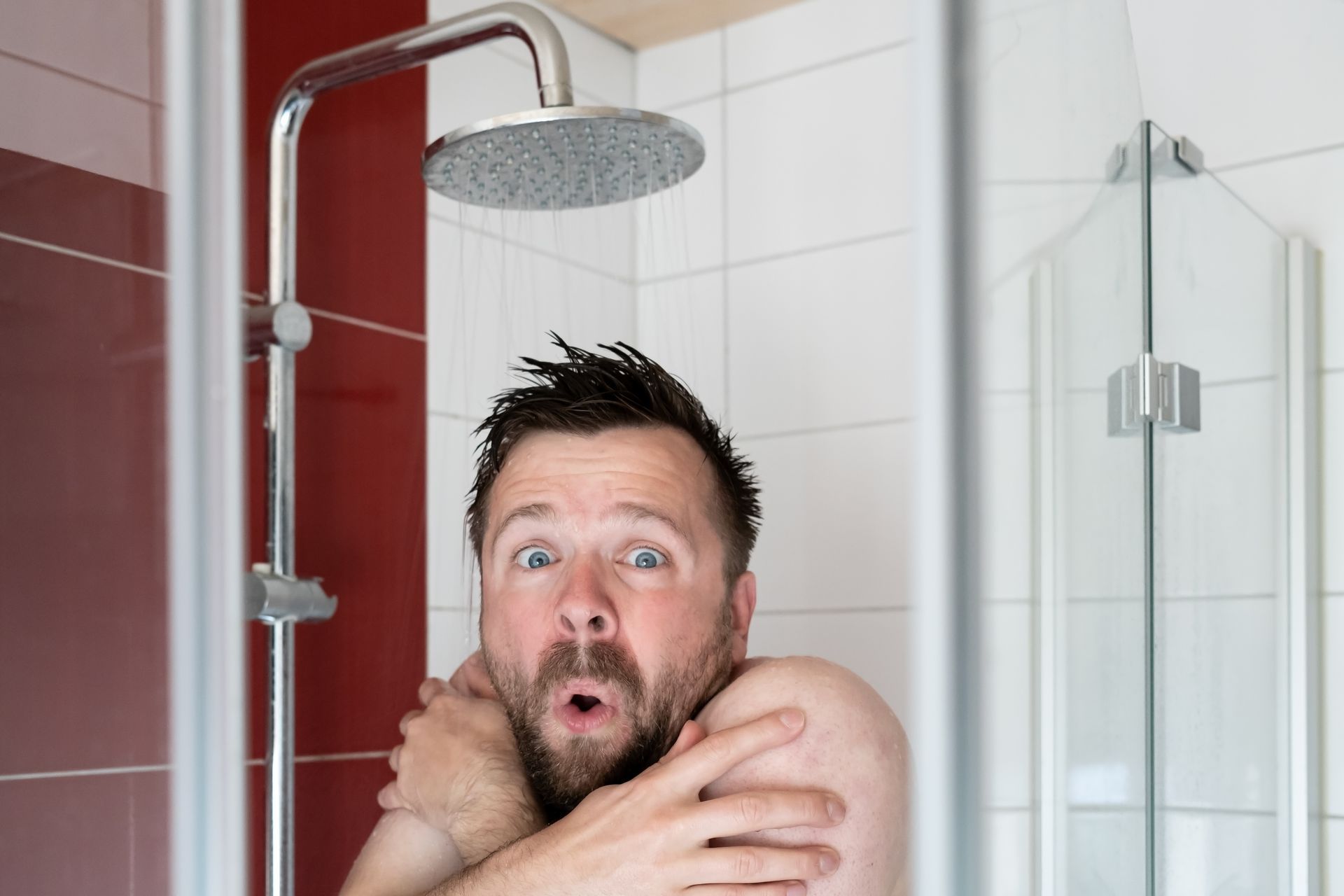 Man in shower with startled expression, arms crossed, under cold water.