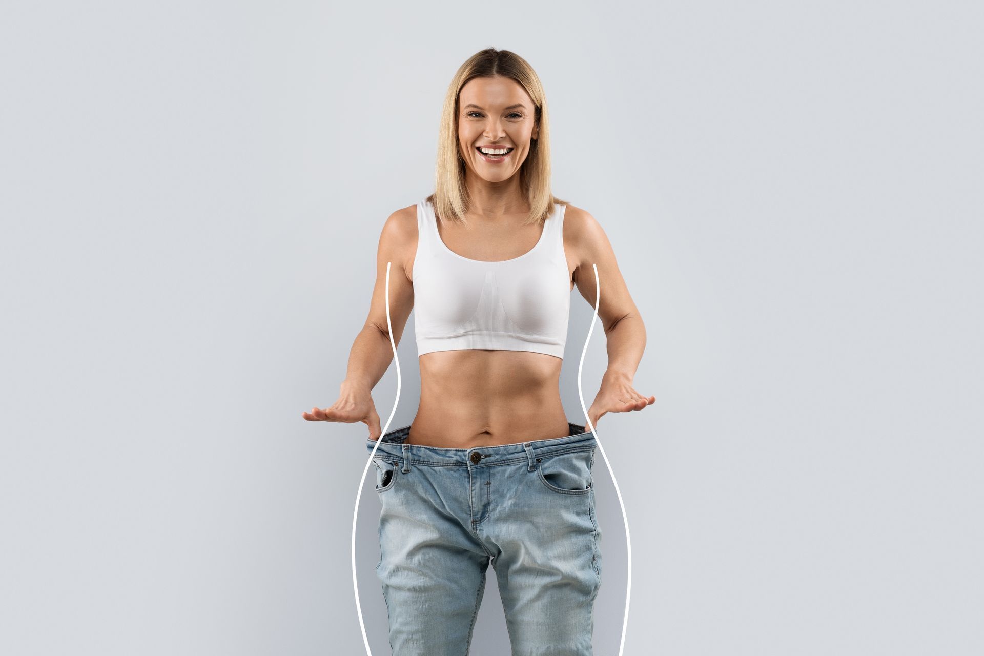 Woman in oversized jeans, showing weight loss, smiling. White top, light background.