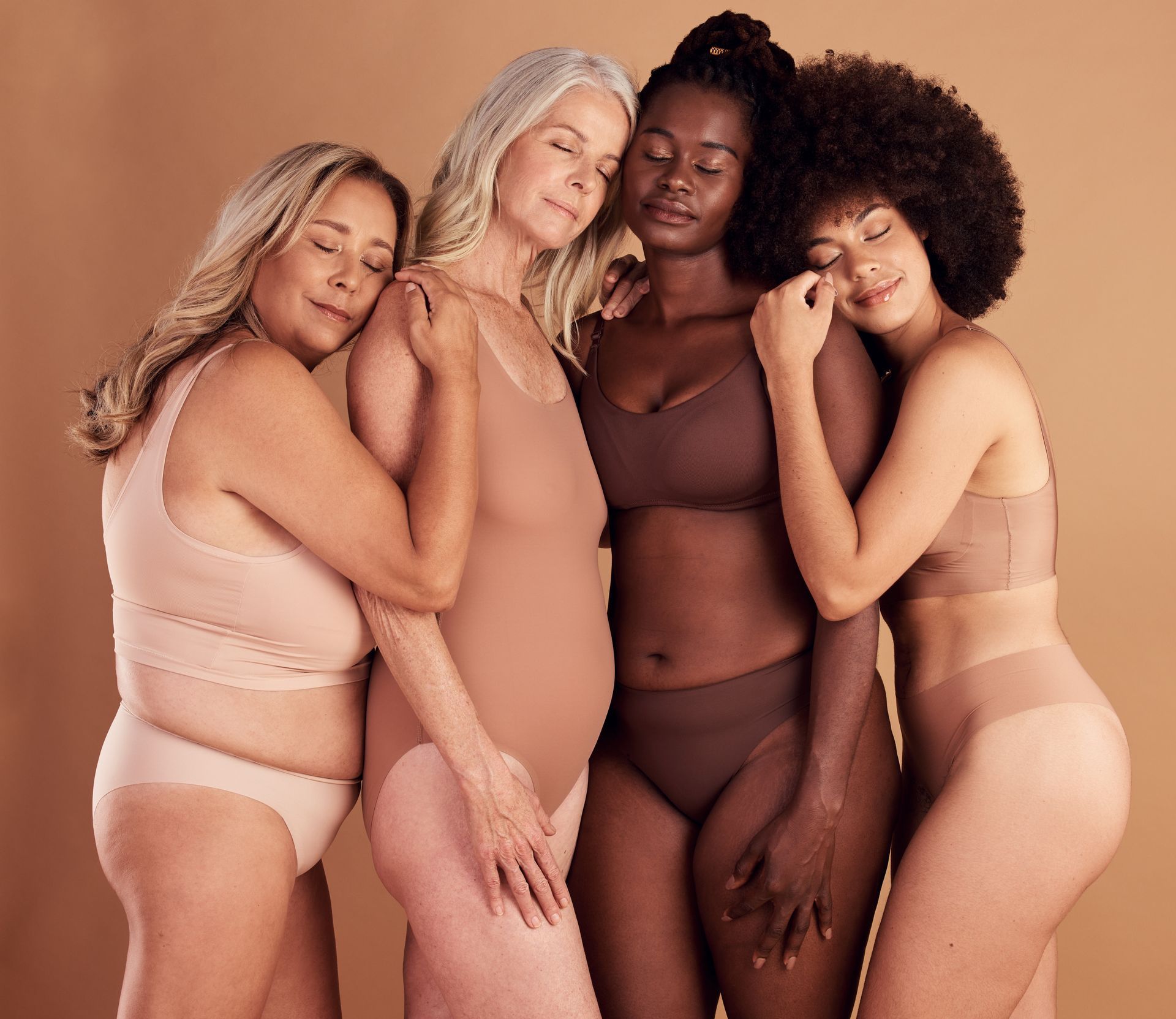 Four people in neutral-toned lingerie embrace, eyes closed, against a beige background.