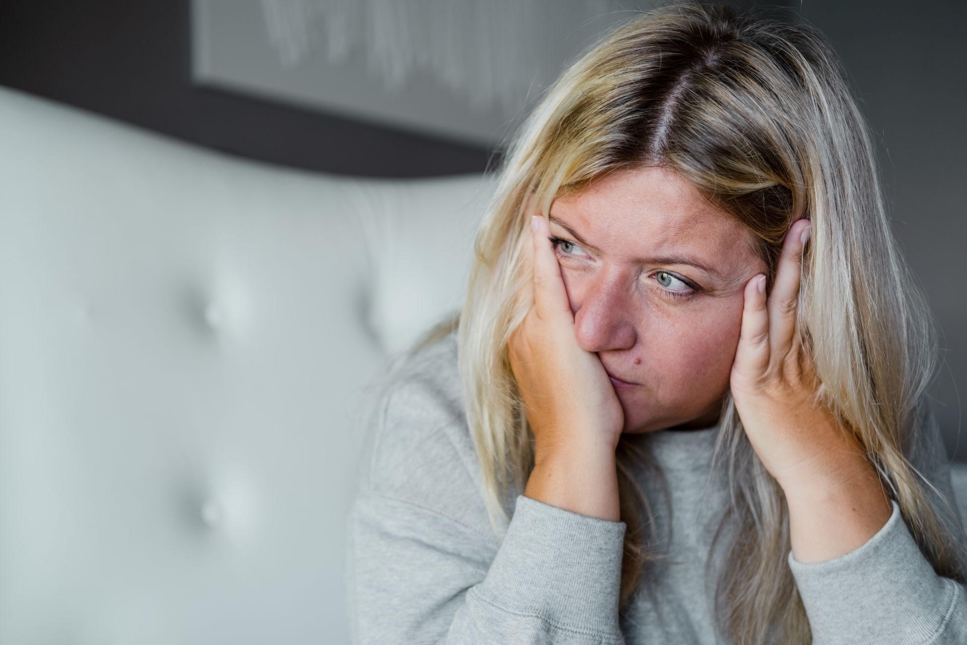 Woman with blonde hair, hands on face, looking distressed. Gray sweatshirt, white background.