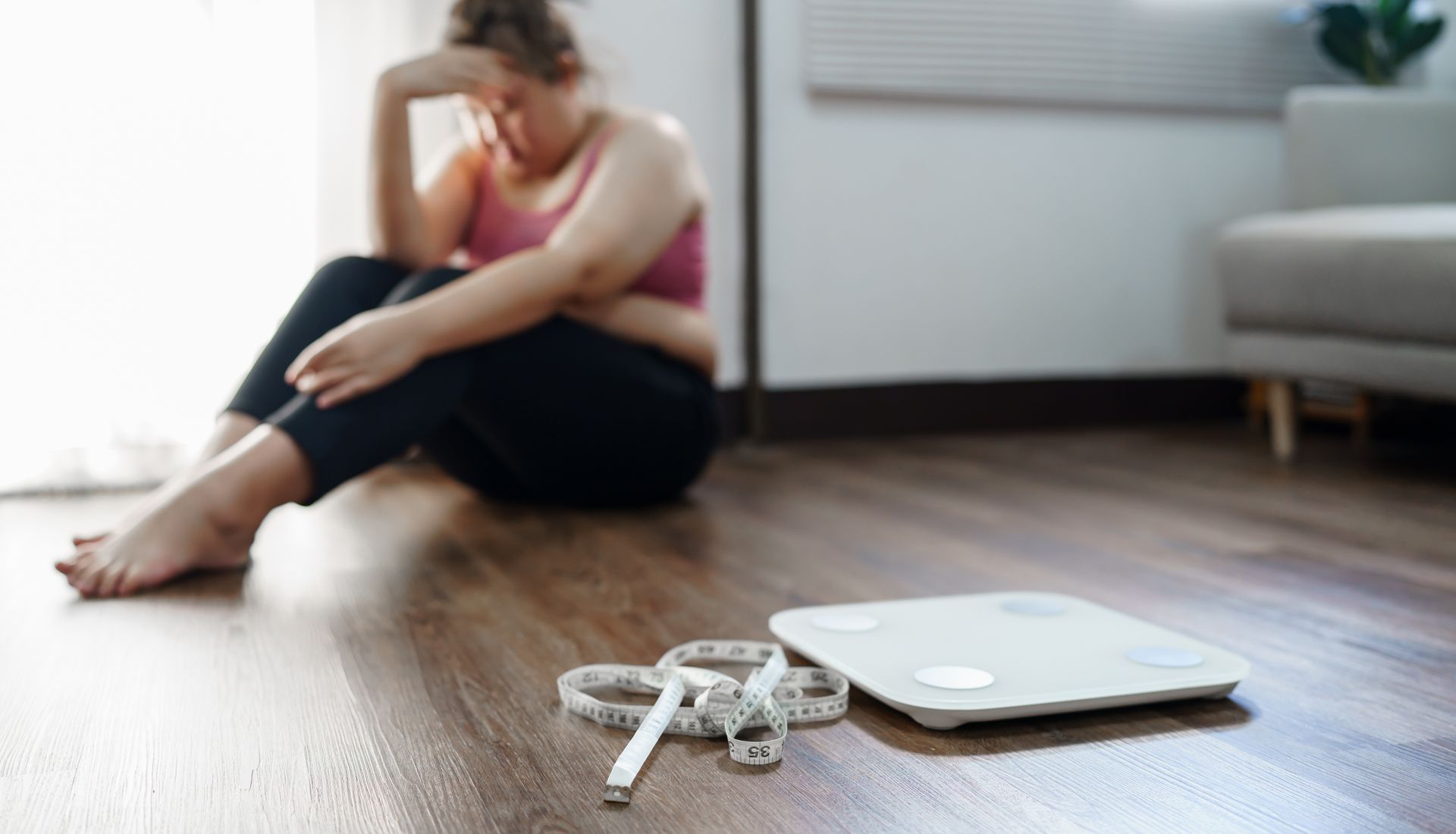 Woman sitting on the floor, looking despondent; scale and measuring tape in foreground.