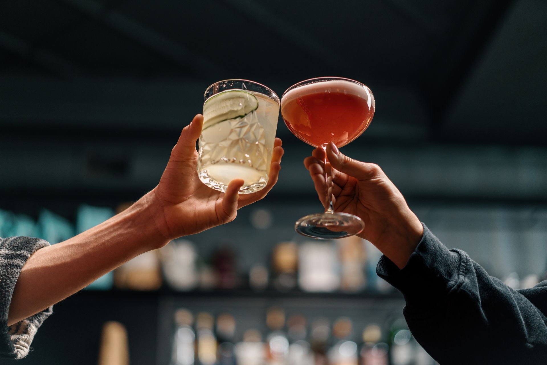 Two people clinking cocktail glasses in a bar. One is clear with cucumber, the other is pink.