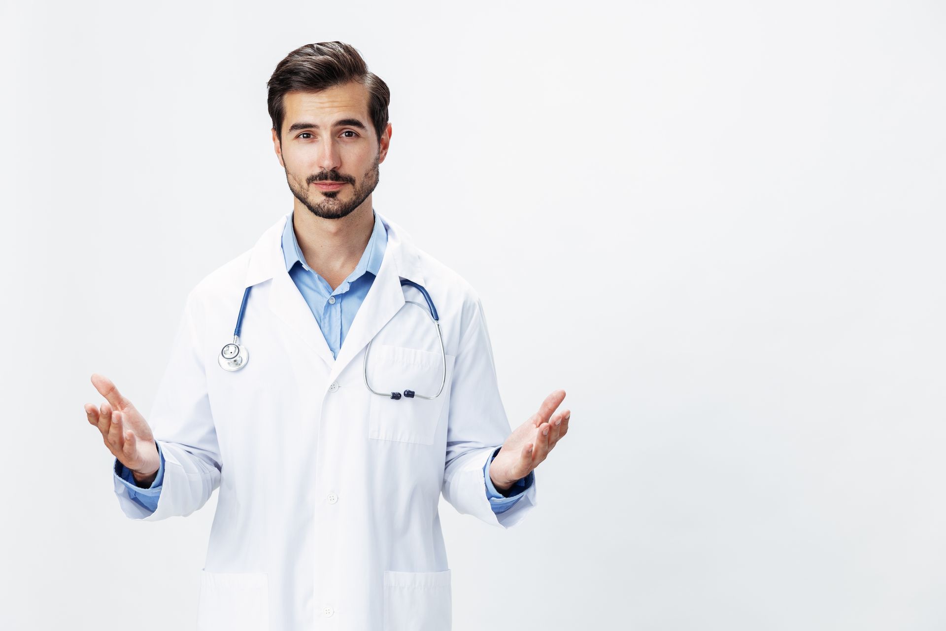 Doctor with stethoscope and white coat gestures with open hands, white background.