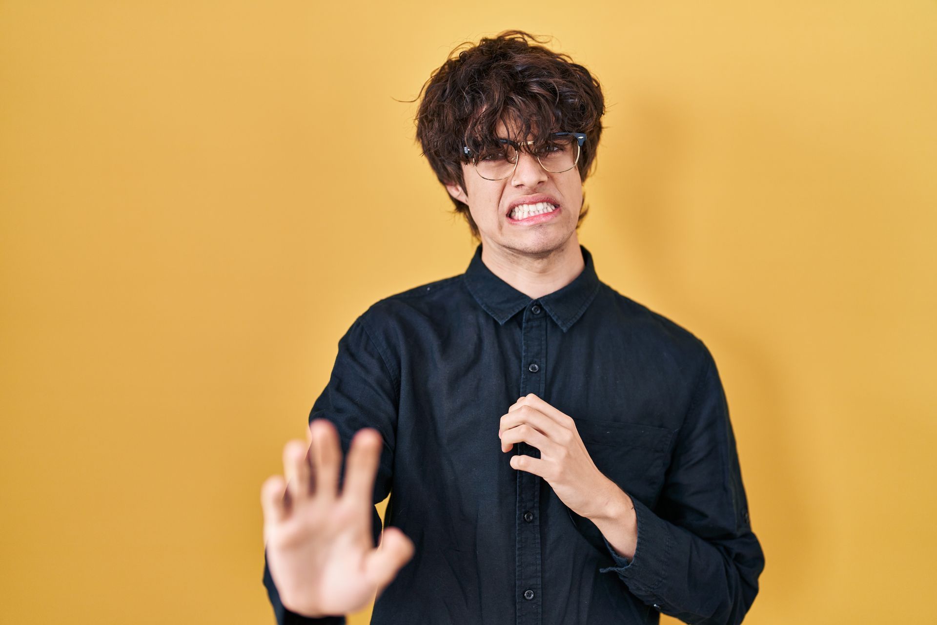 Man in black shirt with disgusted expression, holding out hand, yellow background.