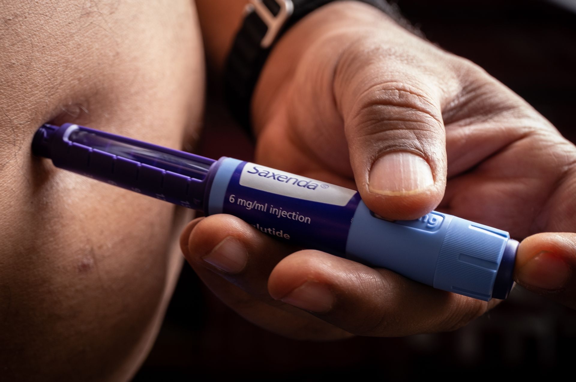 Hand injecting medication with a purple and blue pen injector into an arm.