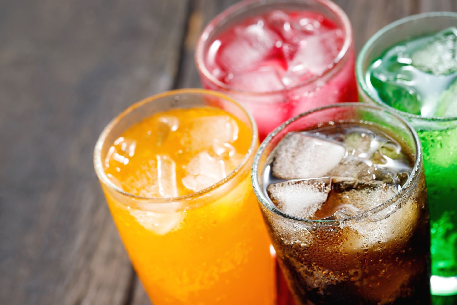 Four colorful sodas in glasses with ice cubes. Orange, red, brown, and green drinks on wooden surface.