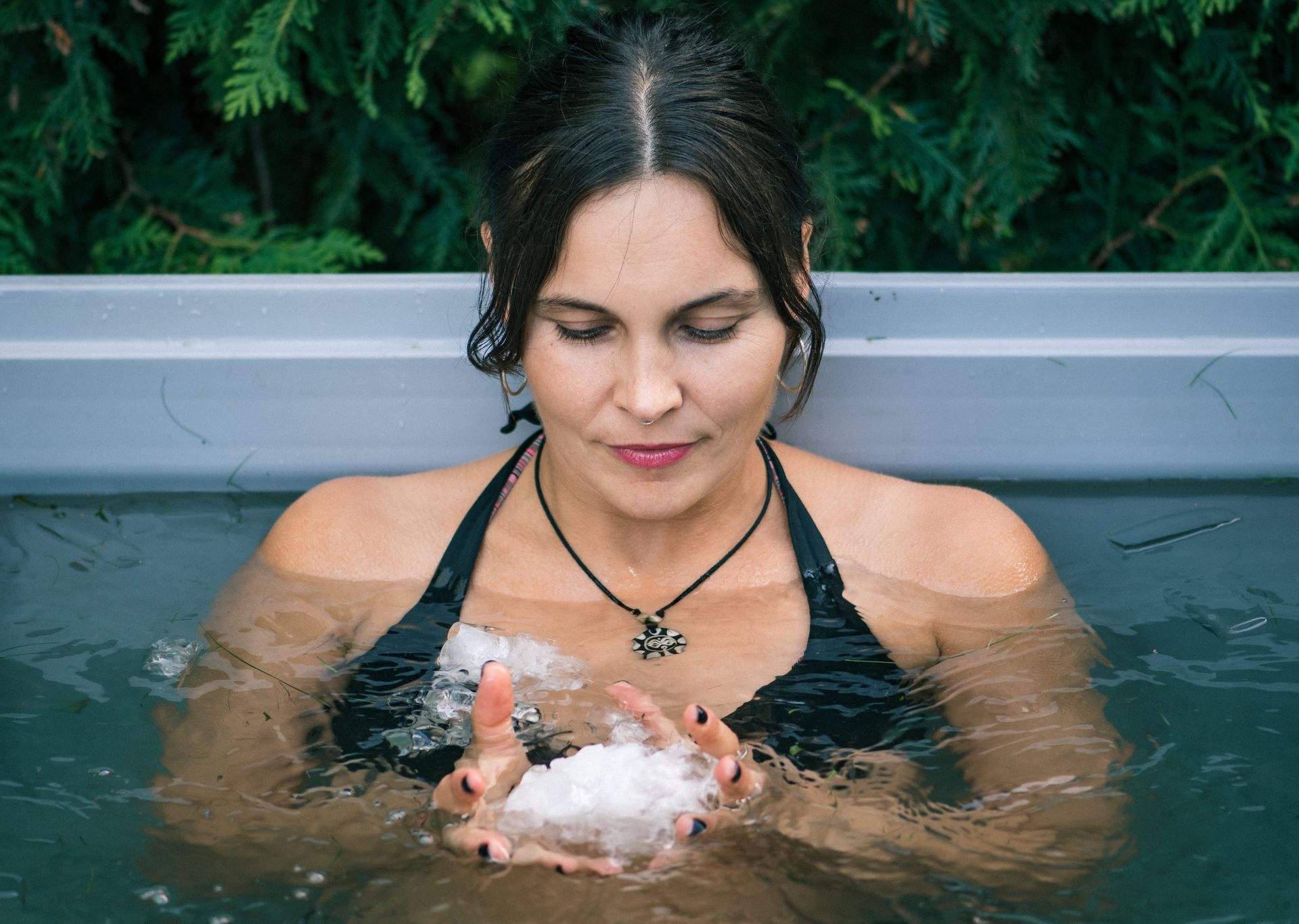 Woman in black swimsuit in a cold plunge pool holding ice, serene expression. Green foliage in background.