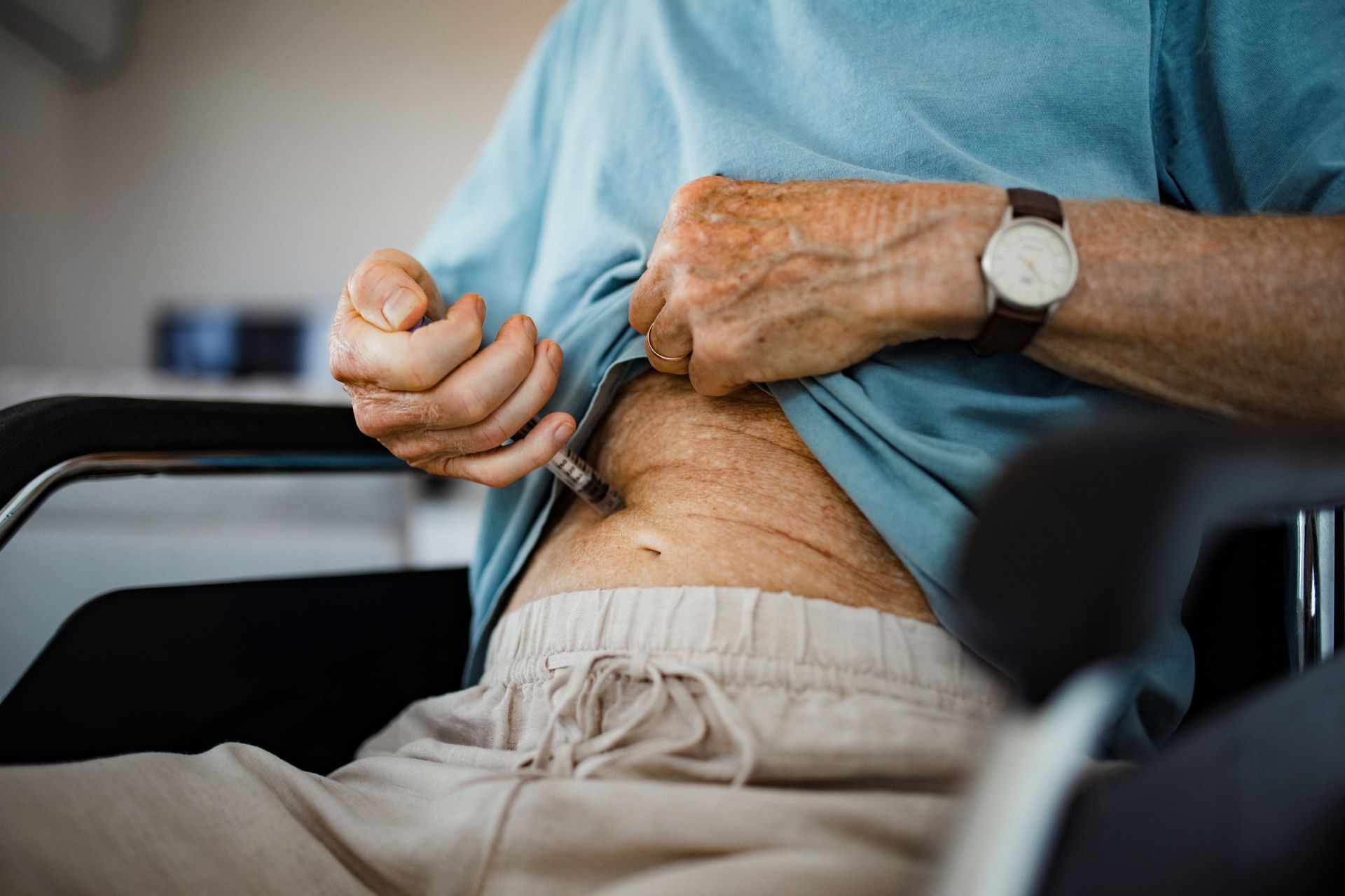 Person injecting medicine into abdomen. Wearing a watch and a blue shirt, seated in a chair.
