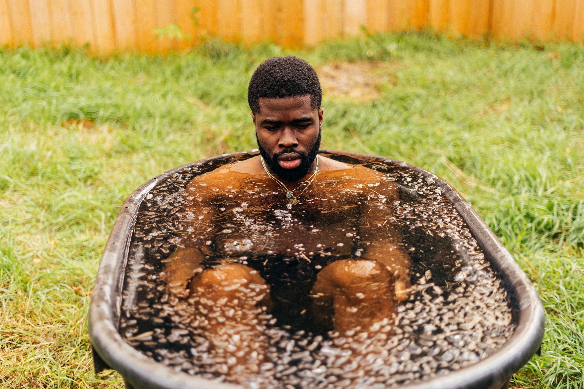 Man sits in a cold plunge tub, outdoors in a yard. Brown-skinned, in black tub with bubbles.