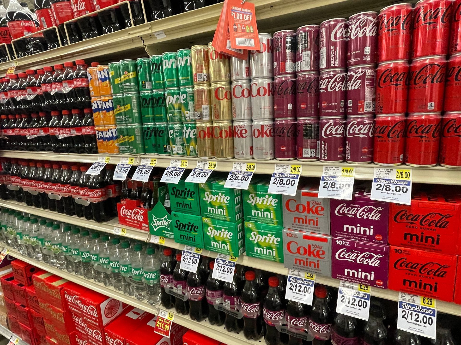 Shelves of various soft drinks, including Coke, Sprite, and Fanta, in cans and bottles at a grocery store.
