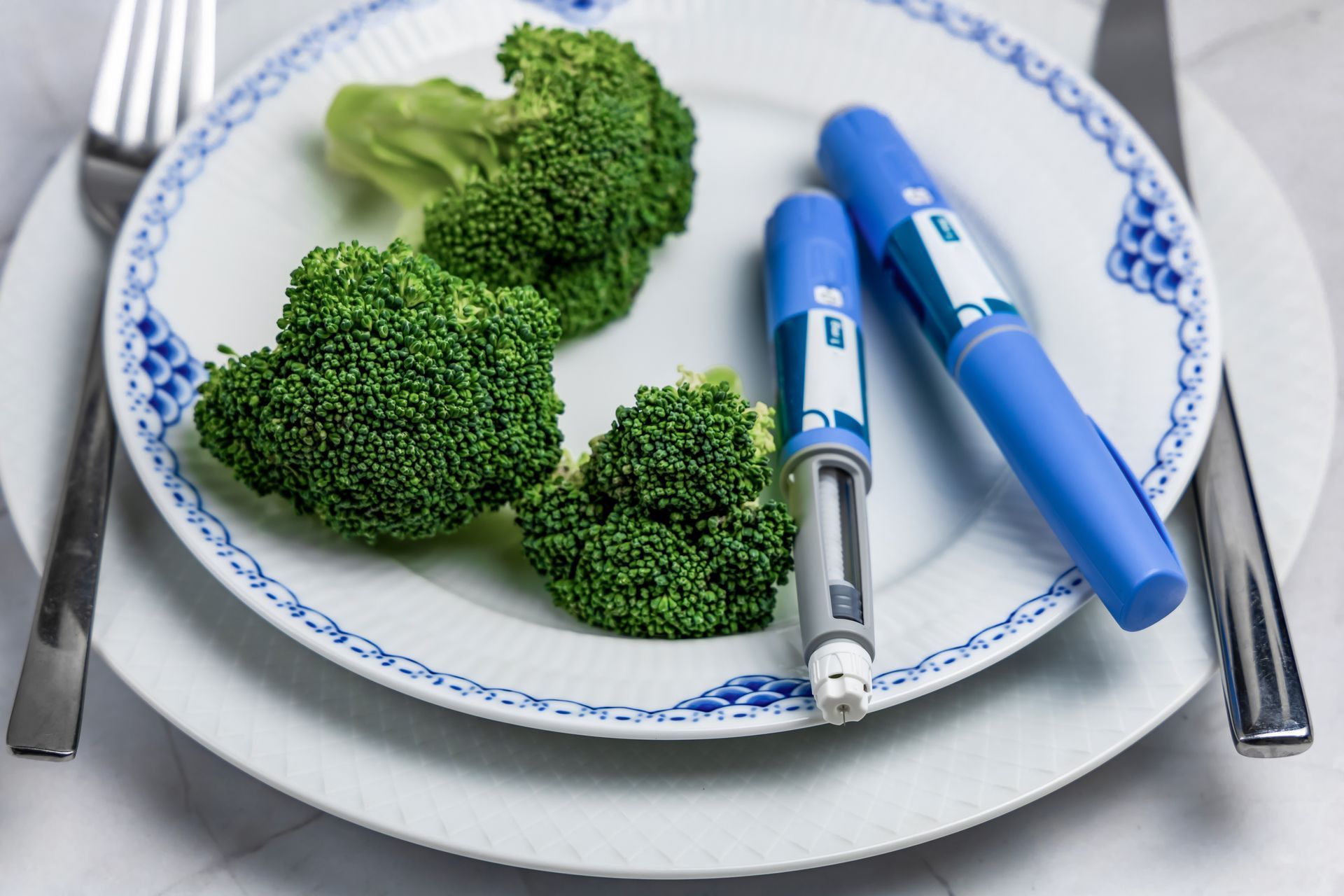Broccoli on a plate with two blue insulin pens, fork and knife on either side.