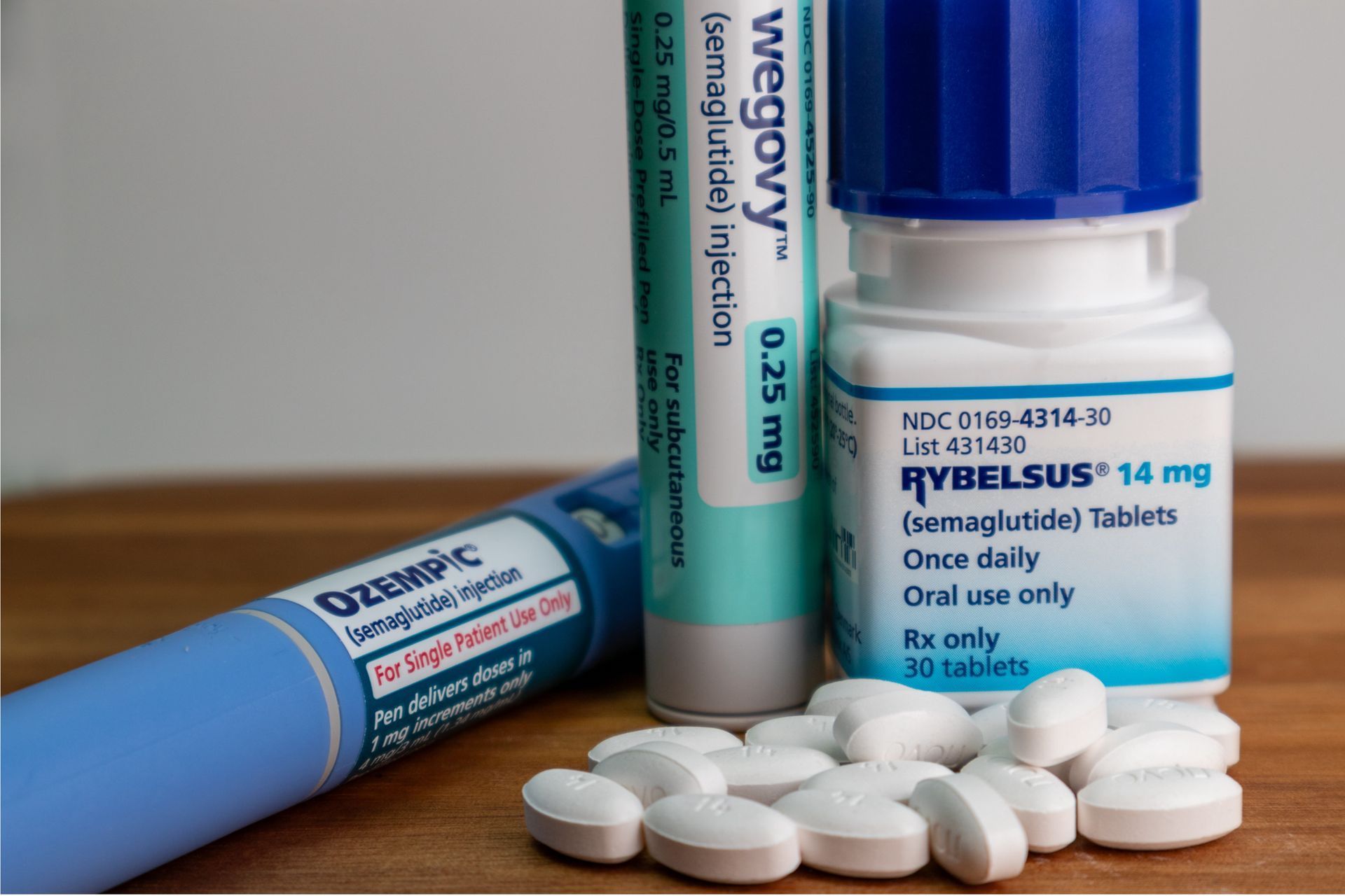 Three prescription medications: Ozempic pen, Wegovy pen, and Rybelsus pills in a bottle, on a wooden surface.