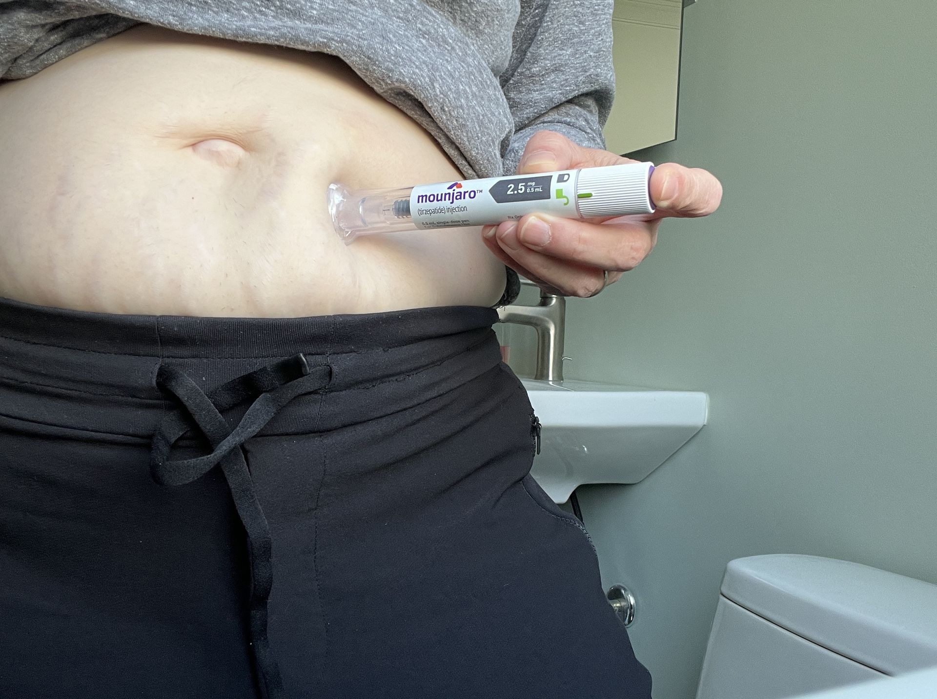 Person injecting medication into abdomen with an injection pen near a bathroom sink and toilet.