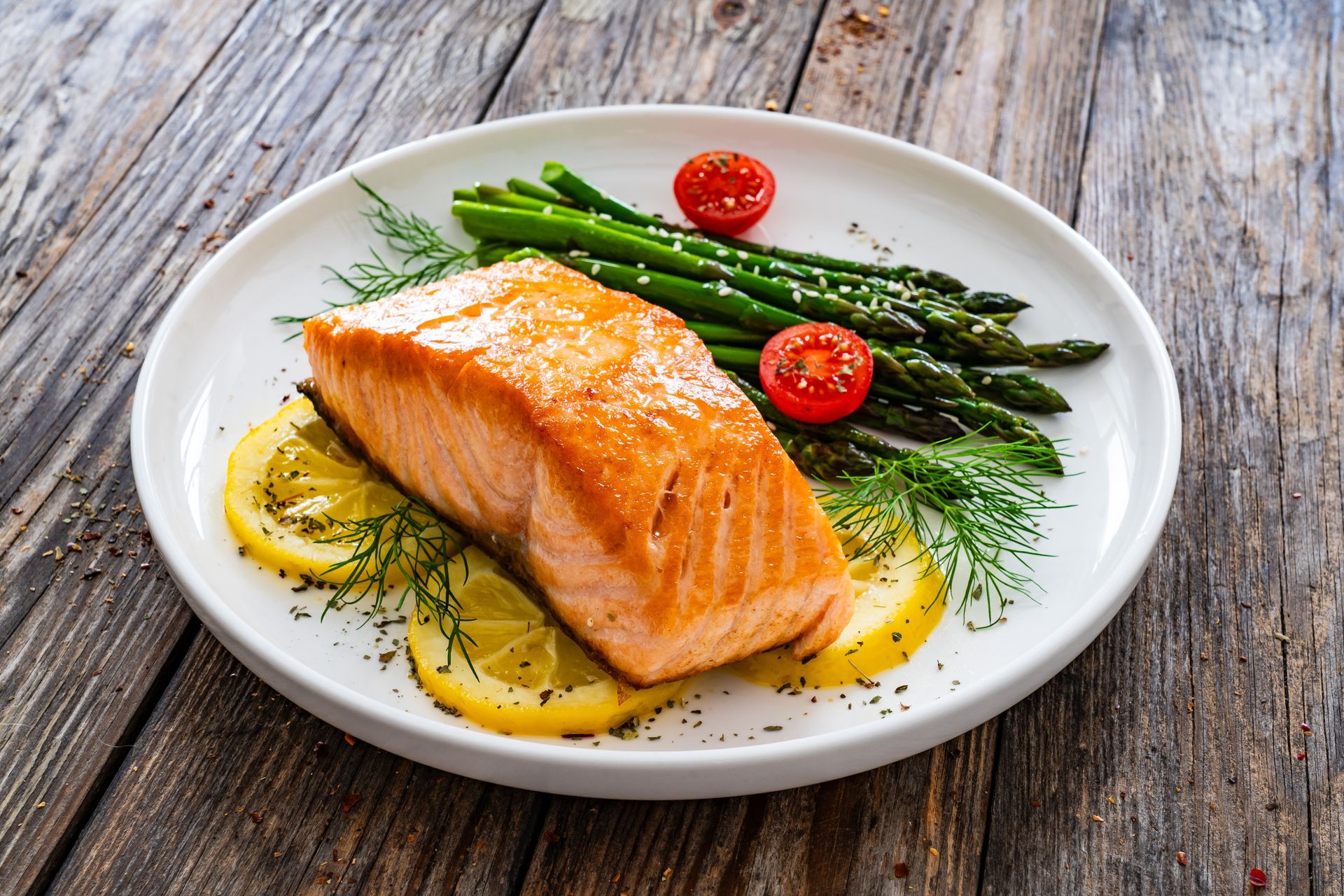 Salmon fillet with asparagus, tomatoes, and lemon slices on a white plate atop a wood table.