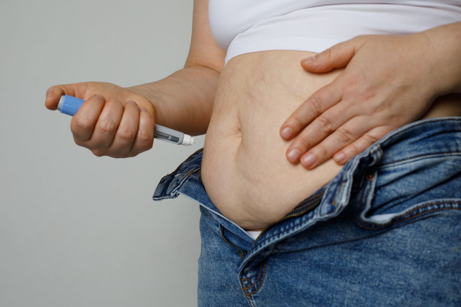 Person injecting medication into abdomen. Unbuttoned jeans, white top, beige skin.