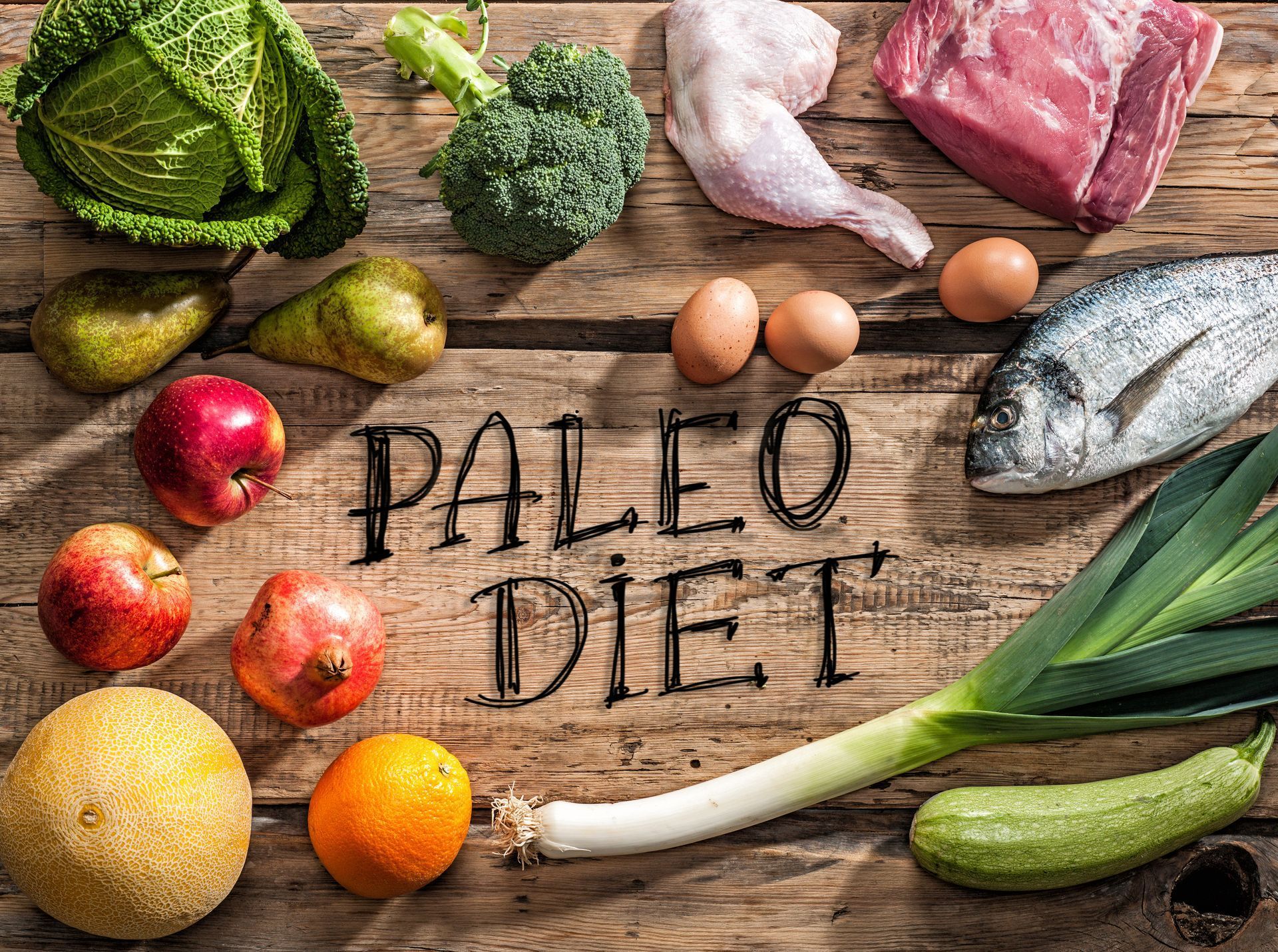 Paleo diet food spread: vegetables, fruits, eggs, fish, and meat on a wooden surface.