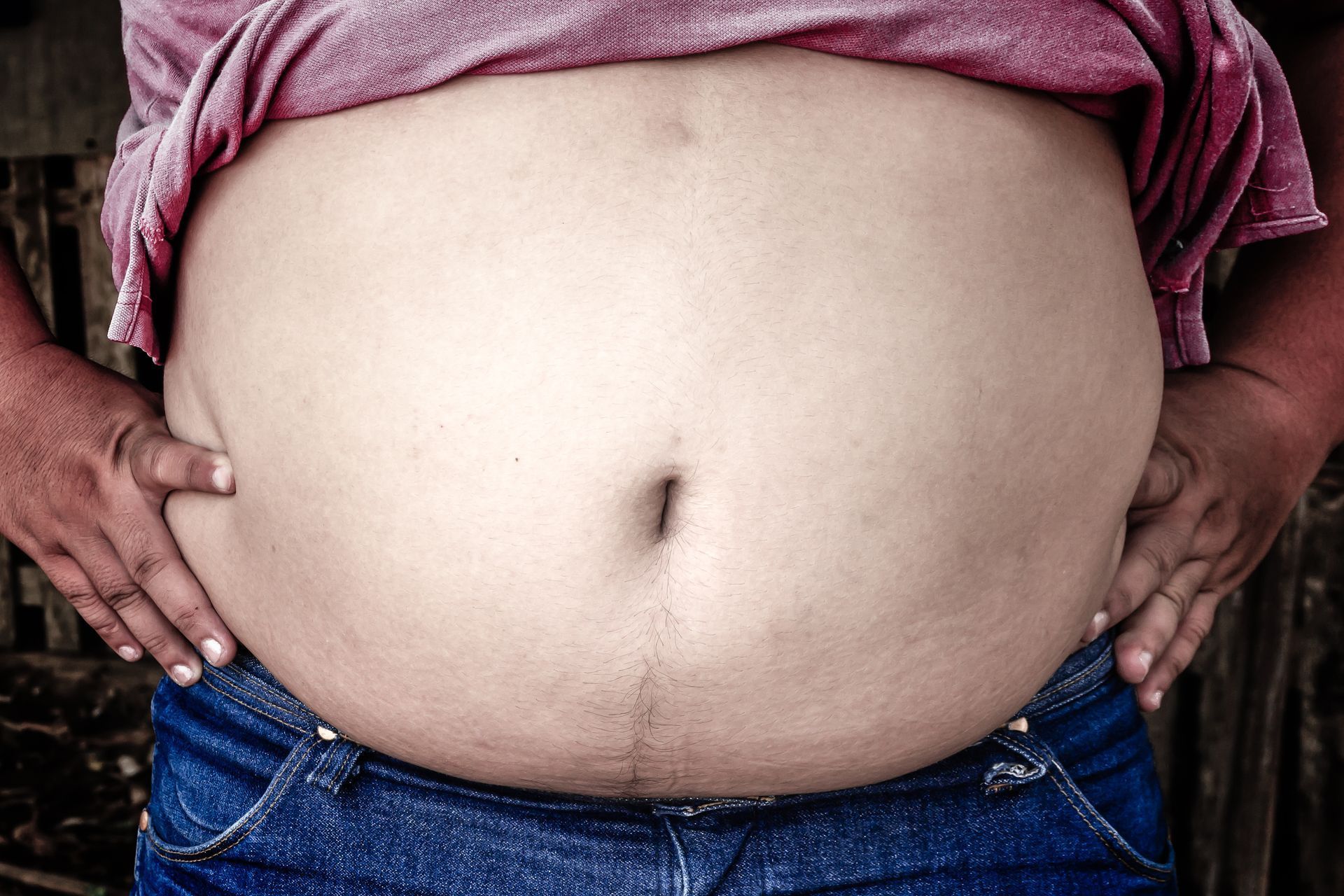 Person's torso with large abdomen, wearing a pink shirt and blue jeans, holding their belly.