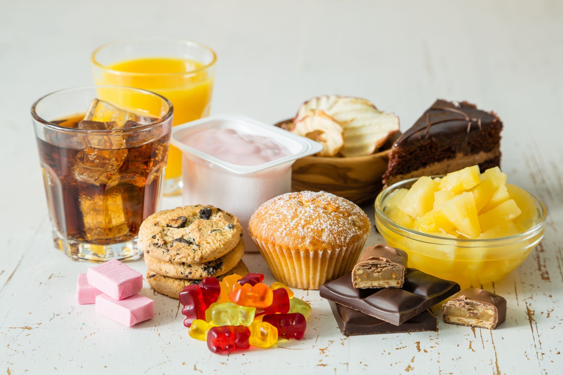 Various sugary foods and drinks on a white surface: cookies, muffins, candy, cake, fruit, soda, juice.
