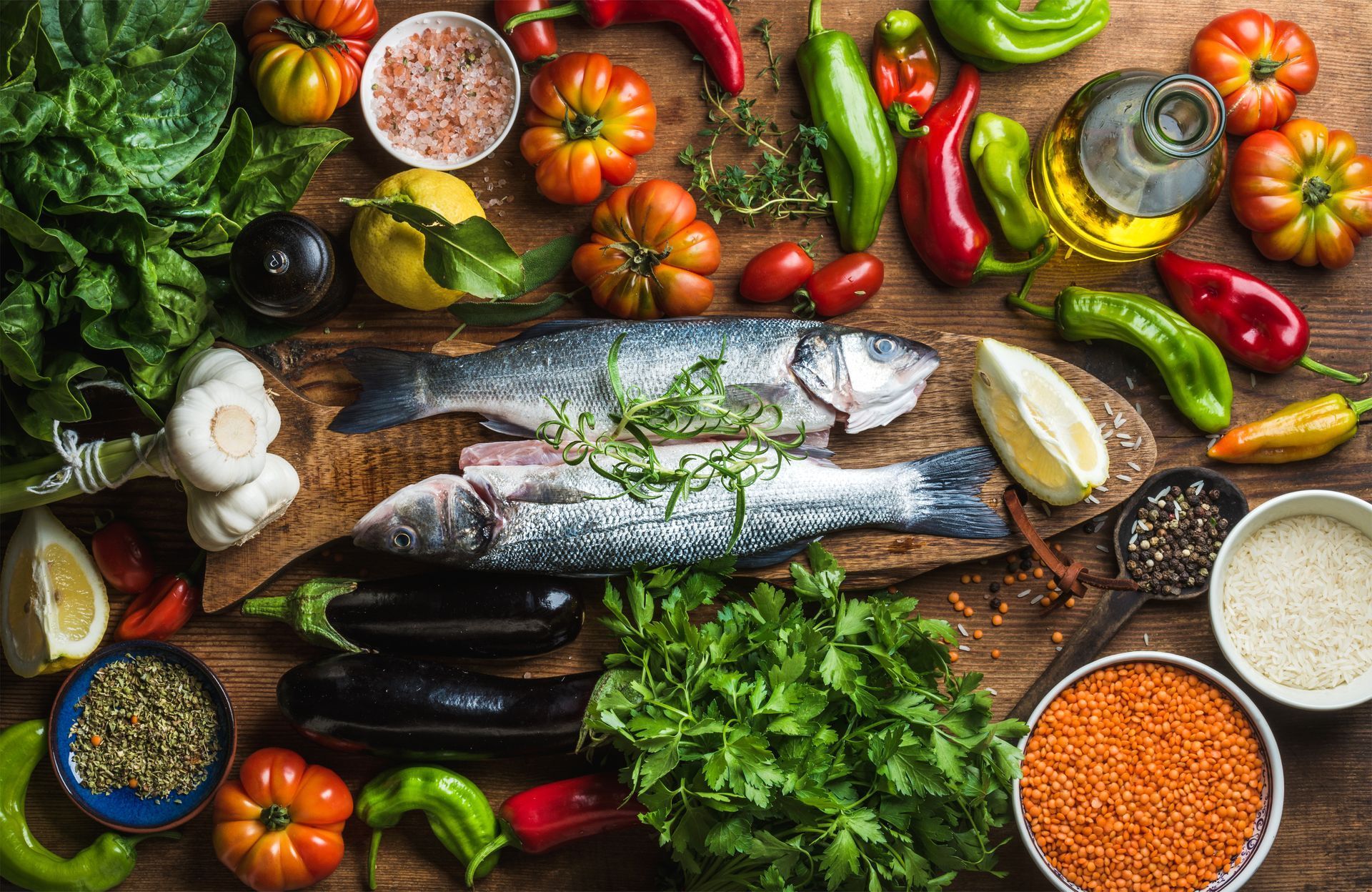 Overhead view of a wooden table with fresh fish, colorful vegetables, spices, and oil.