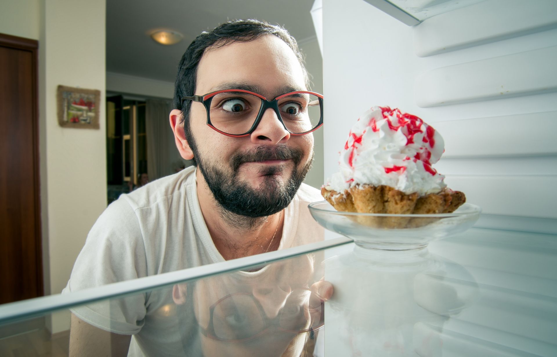 Man with glasses, wide-eyed, peers into refrigerator at a cupcake with white frosting and red sprinkles.