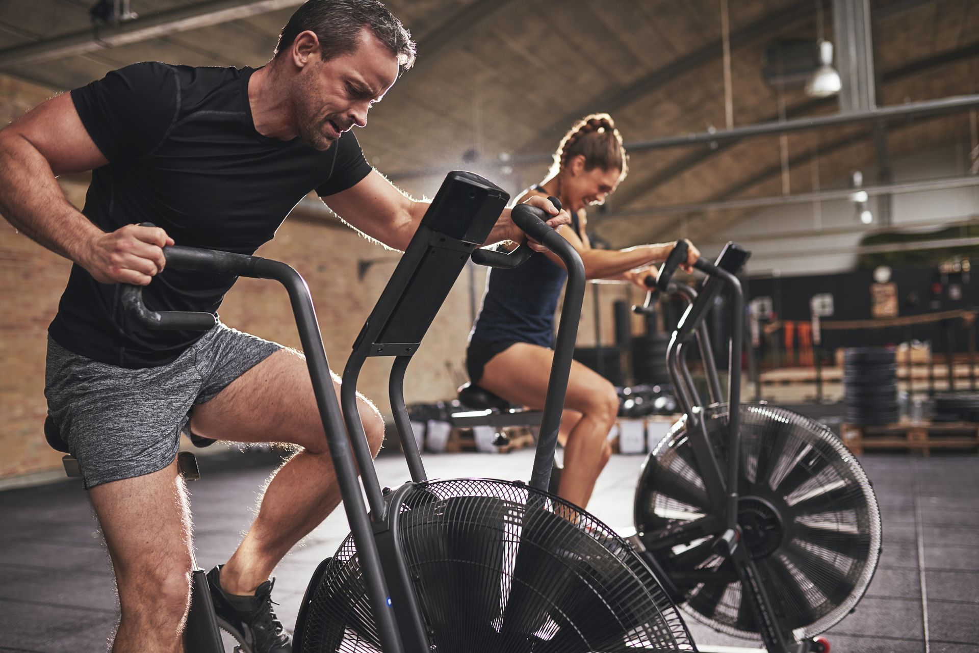 Man and woman exercising on air bikes in a gym, pulling handles and pedaling.