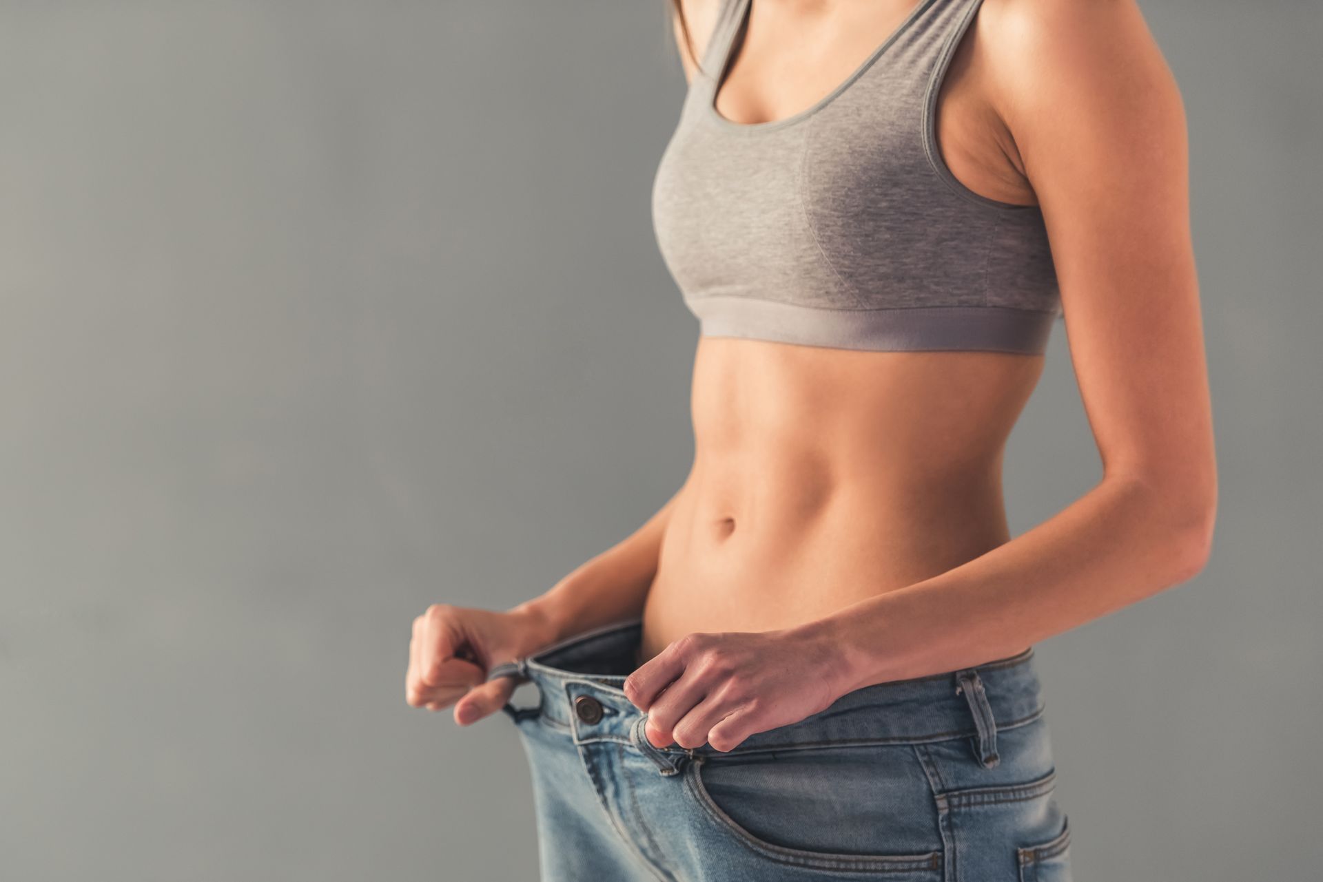 Woman in a sports bra and loose jeans, holding out the waistband, showing weight loss. Gray background.