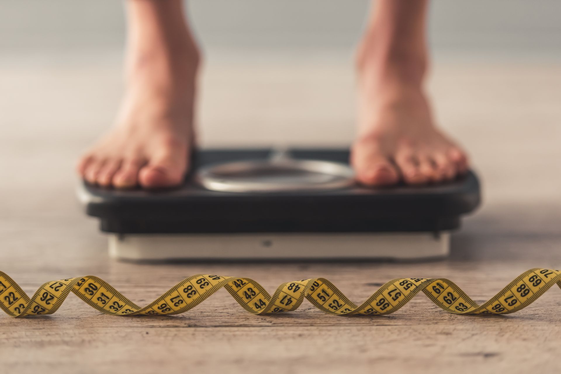 Feet on a scale; yellow measuring tape in front, on wooden floor.