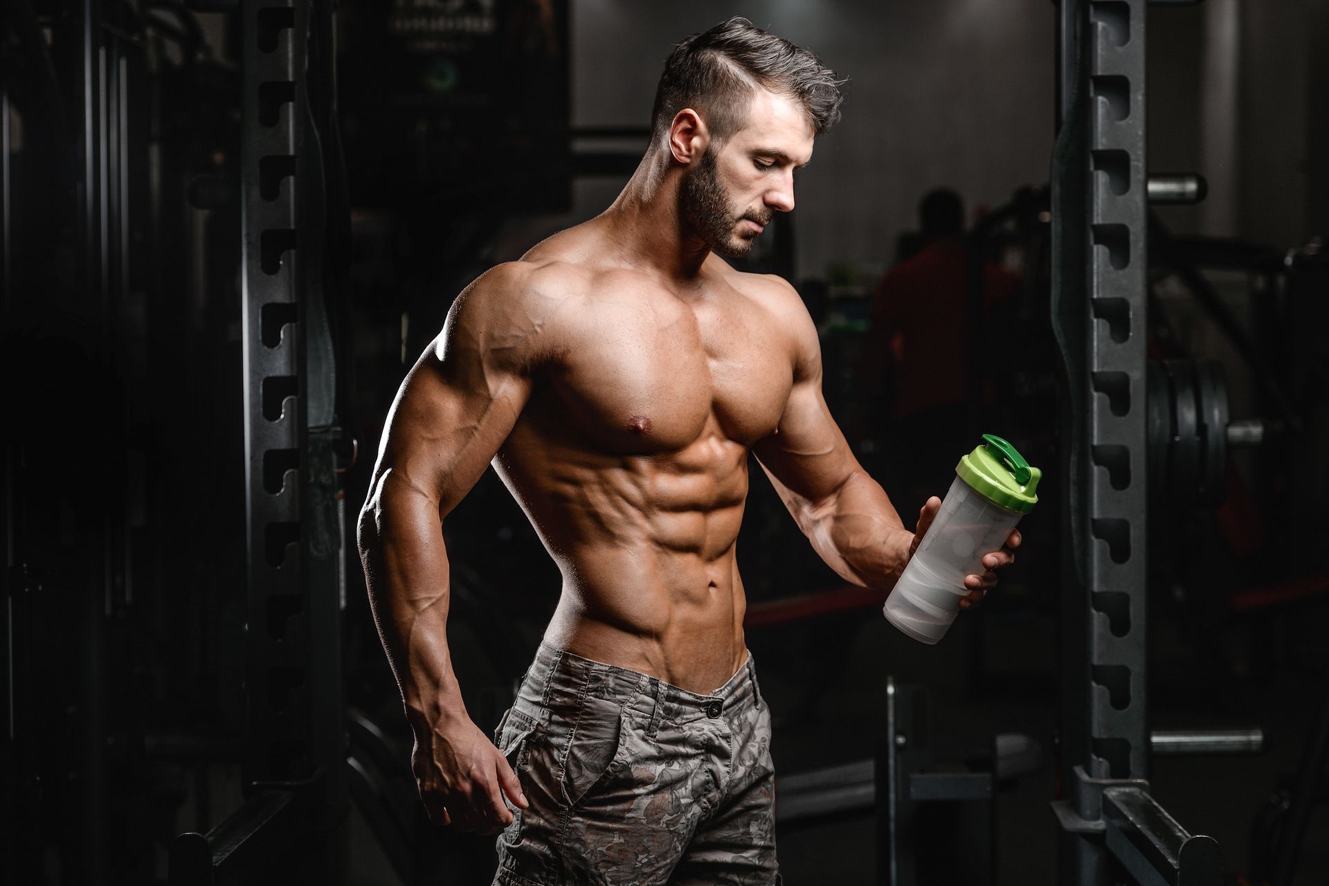 Muscular man in gym holding shaker bottle, looking down. Fitted camo pants, wet skin, dark background.