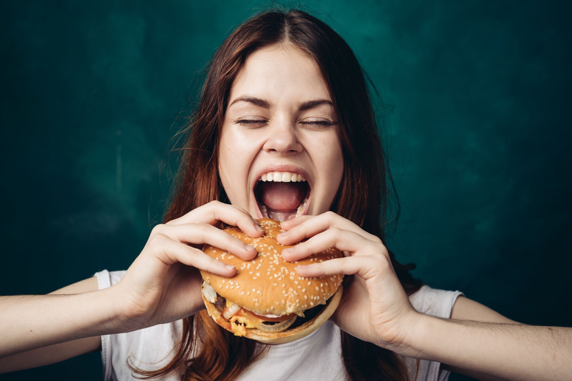Woman taking a large bite of a hamburger, mouth wide open, against a green background.