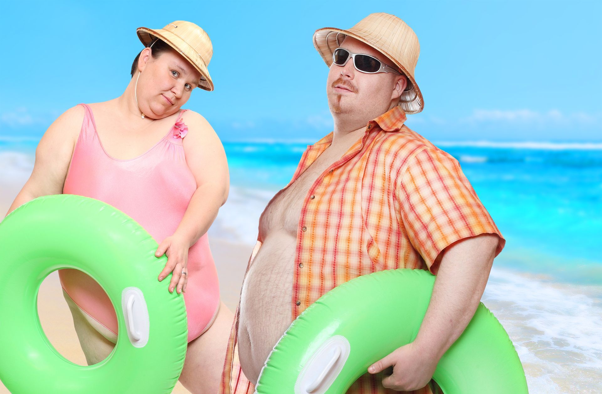 Couple at beach holding inflatable rings; man in sunglasses and open shirt, woman in swimsuit, both wearing hats.