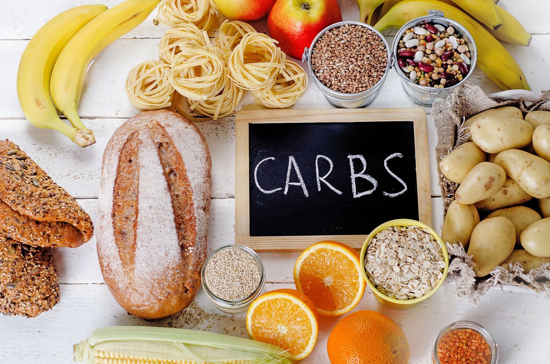 Assorted carbohydrates: bread, pasta, fruit, grains, and potatoes arranged around a chalkboard that says 