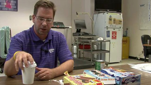 Man in purple shirt holds a coffee cup, sitting at a table with snacks and a computer in the background.
