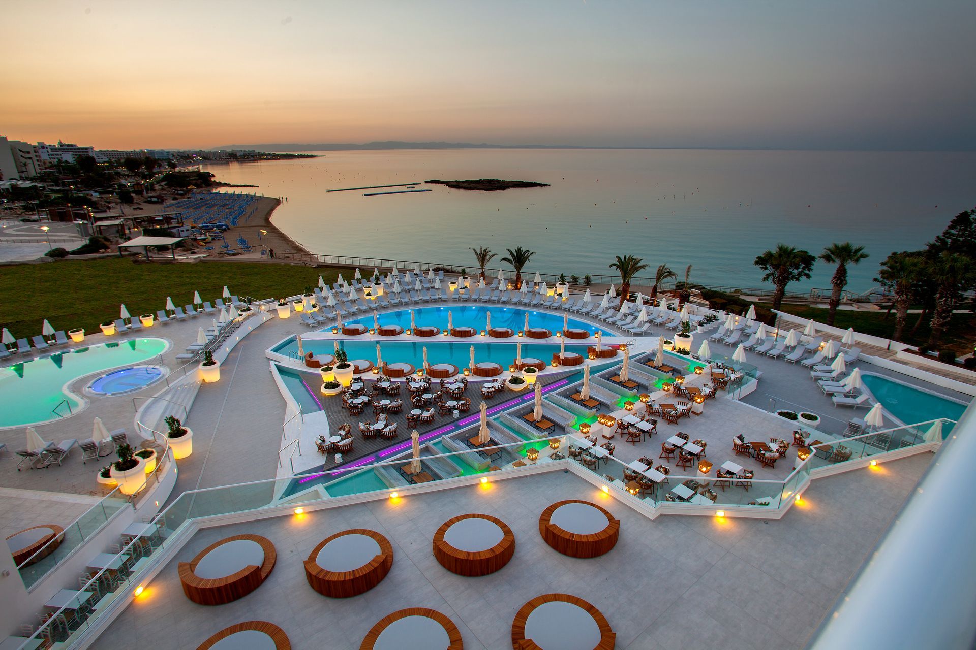 King Jason Protaras pool area overlooking Fig Tree Bay at sunset, with tables, lounge chairs, and ocean view.