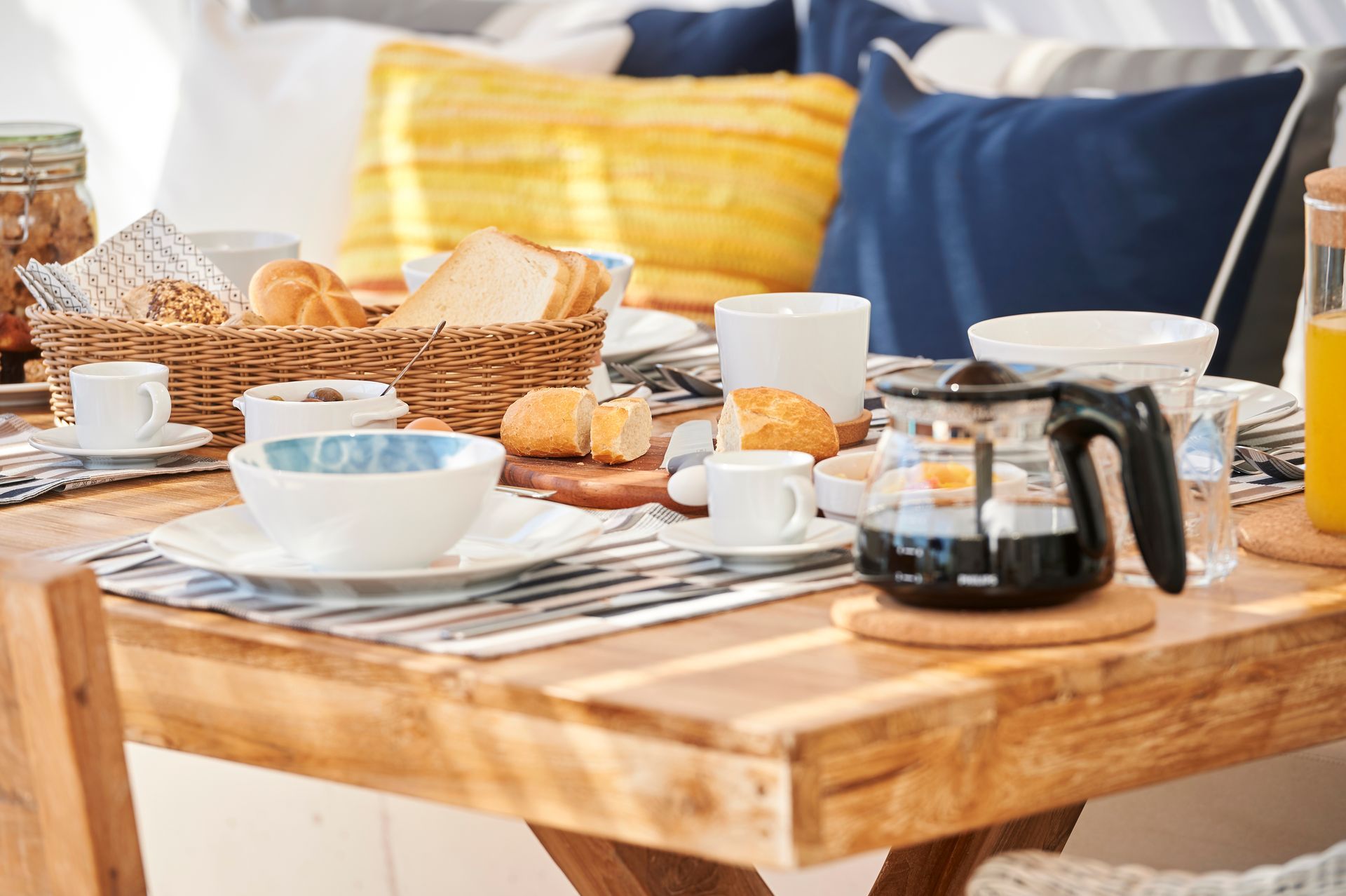 Breakfast table set with coffee, bread, and pastries; blue and yellow pillows in background.