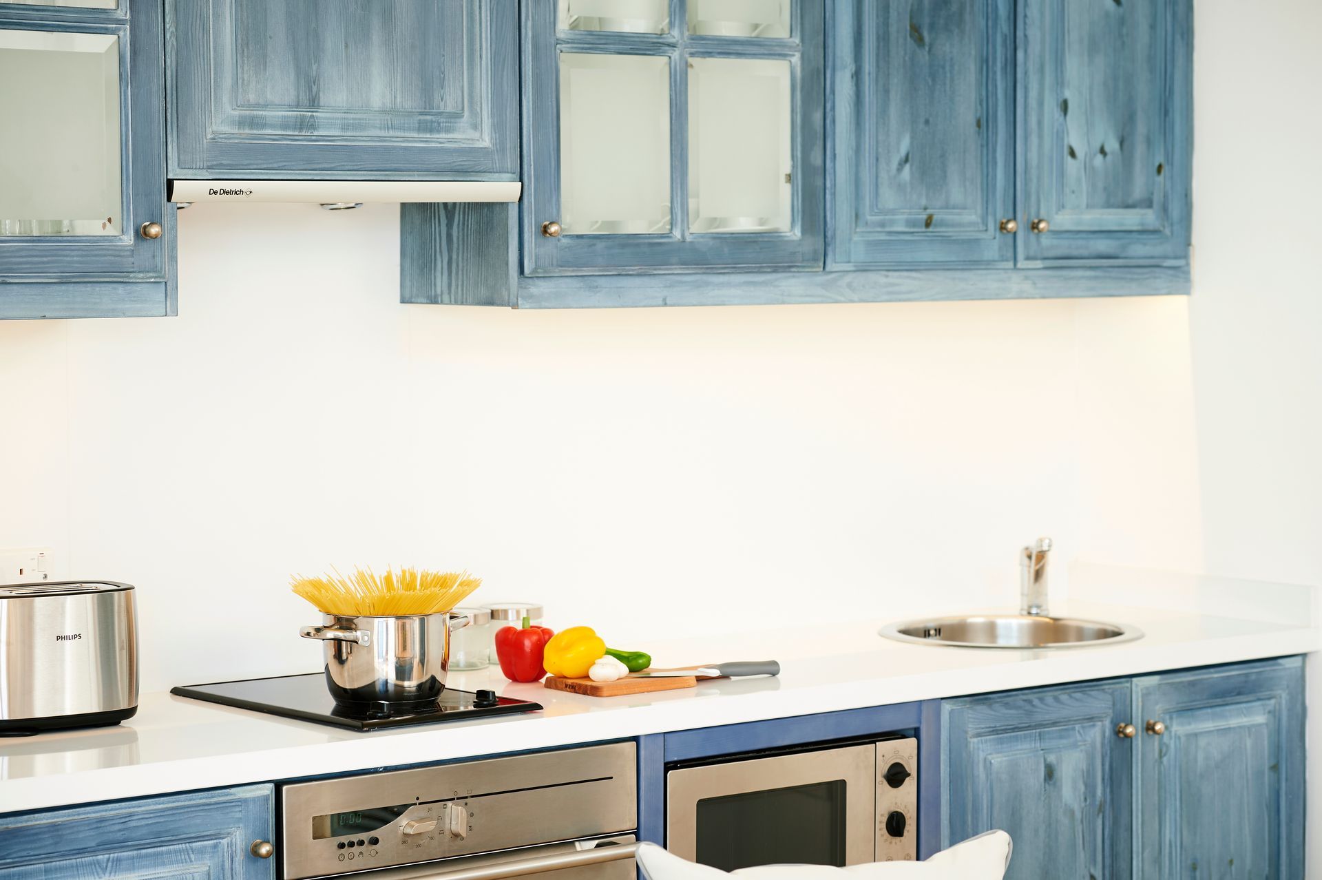 Kitchen with blue cabinets, white countertops, stove with pot, vegetables, and small sink.