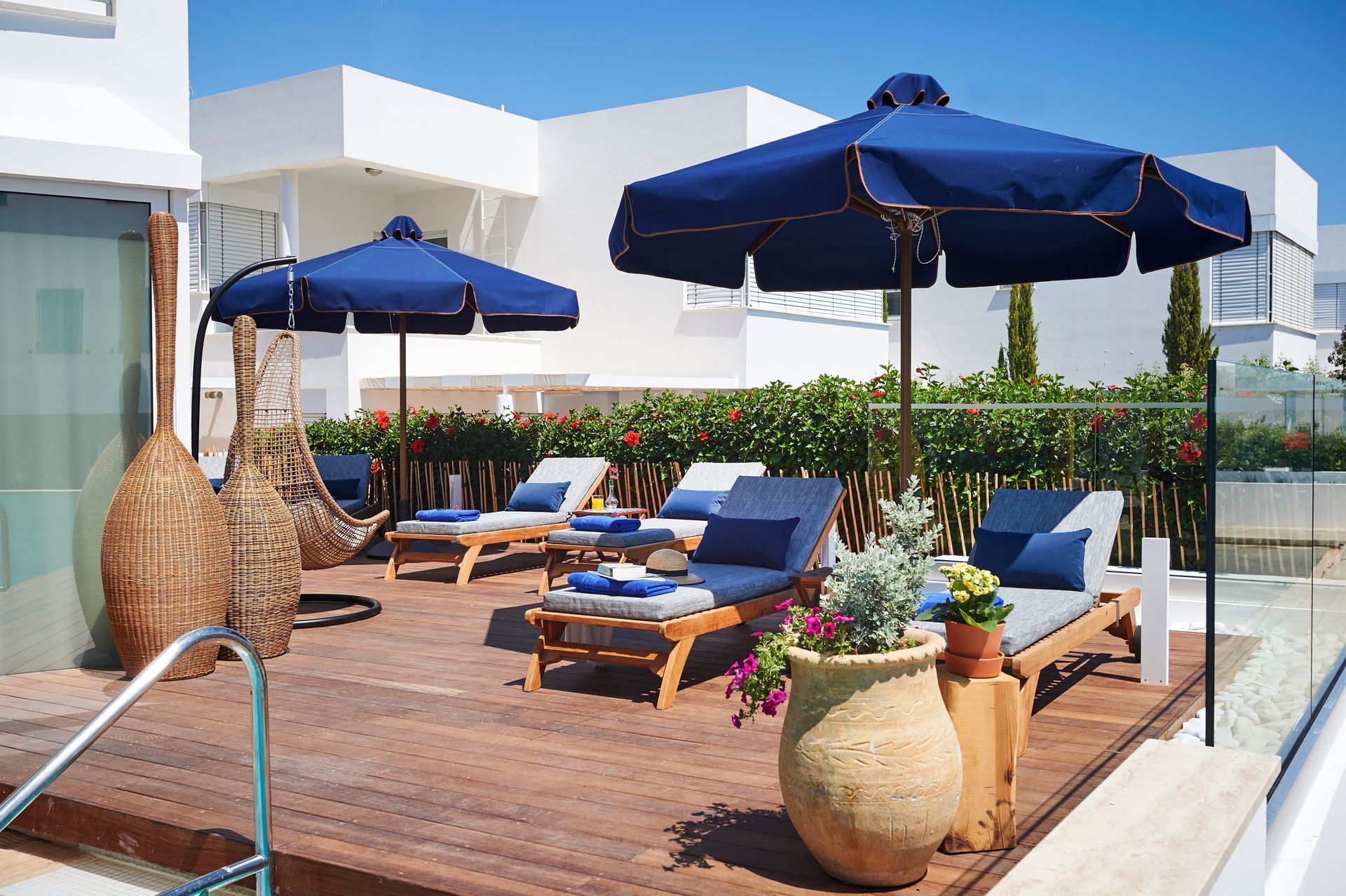 Rooftop deck with lounge chairs, blue umbrellas, and a large decorative pot under a bright blue sky.