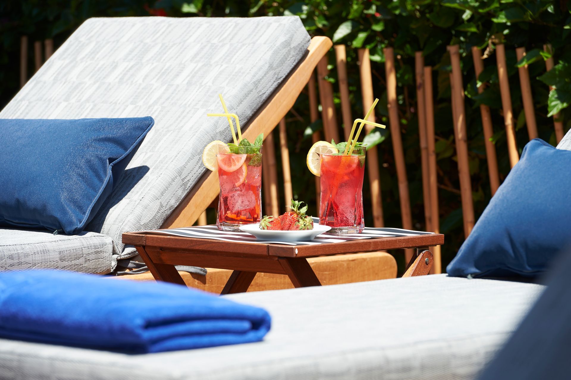 Two red cocktails with lemon, mint, and straws on a tray between lounge chairs by a fence.