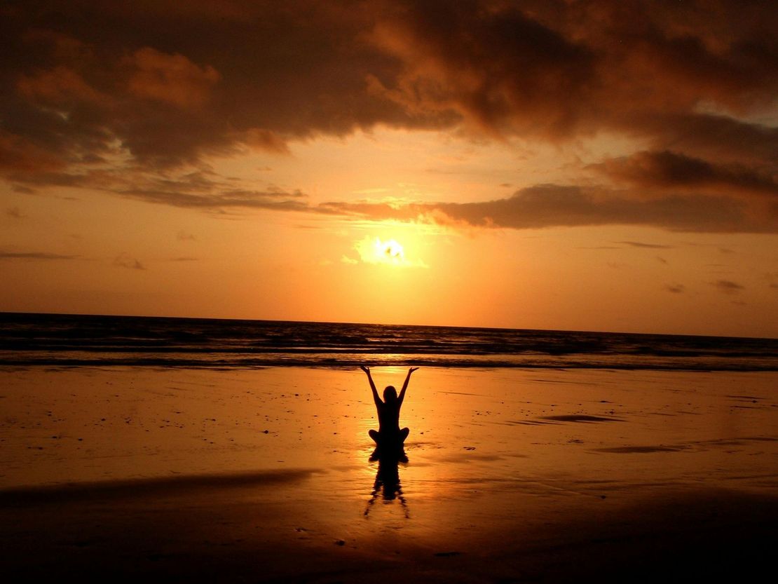 A person is standing on a beach at sunset with their arms in the air