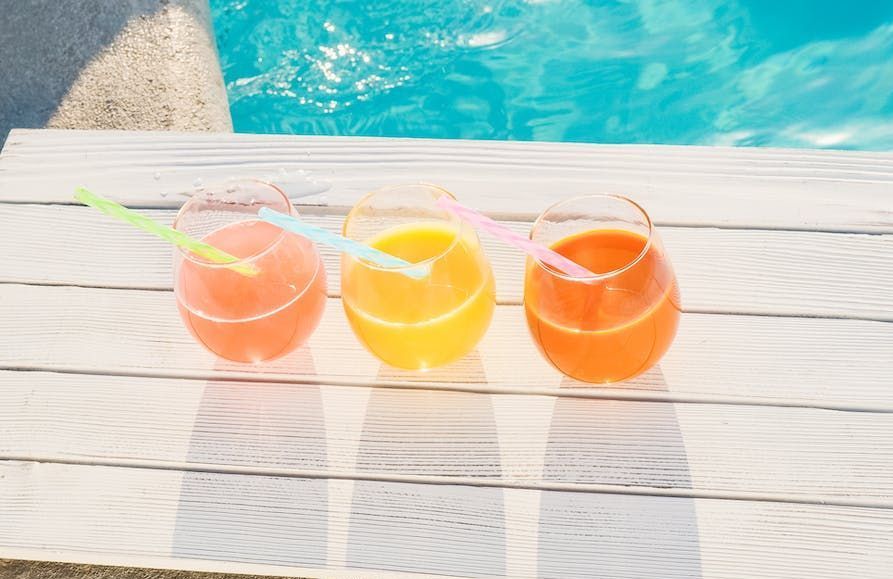 Three glasses of juice are sitting on a wooden table next to a swimming pool.