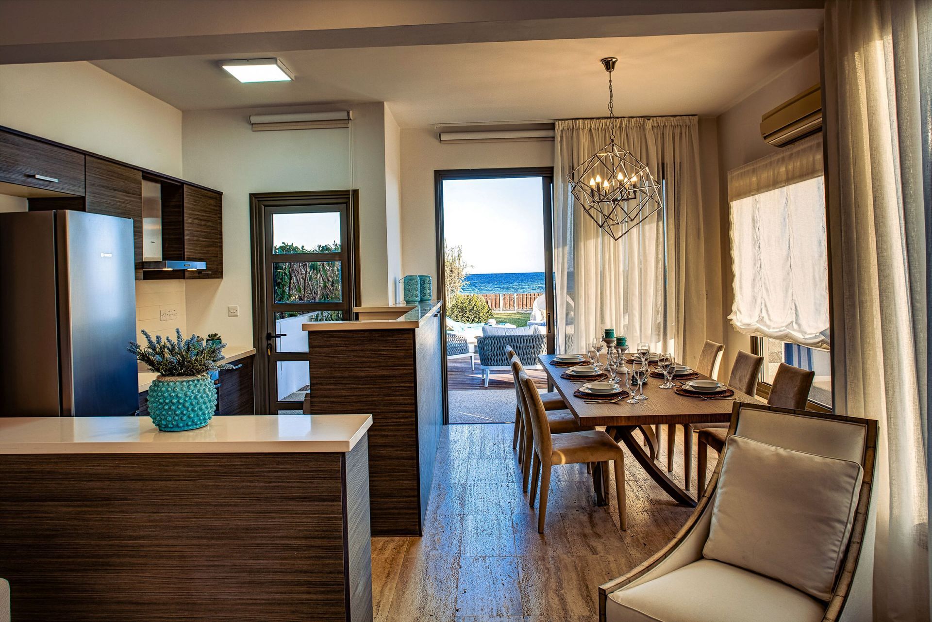 Interior view: open kitchen and dining area with sea view, wooden table and chairs, chandelier.