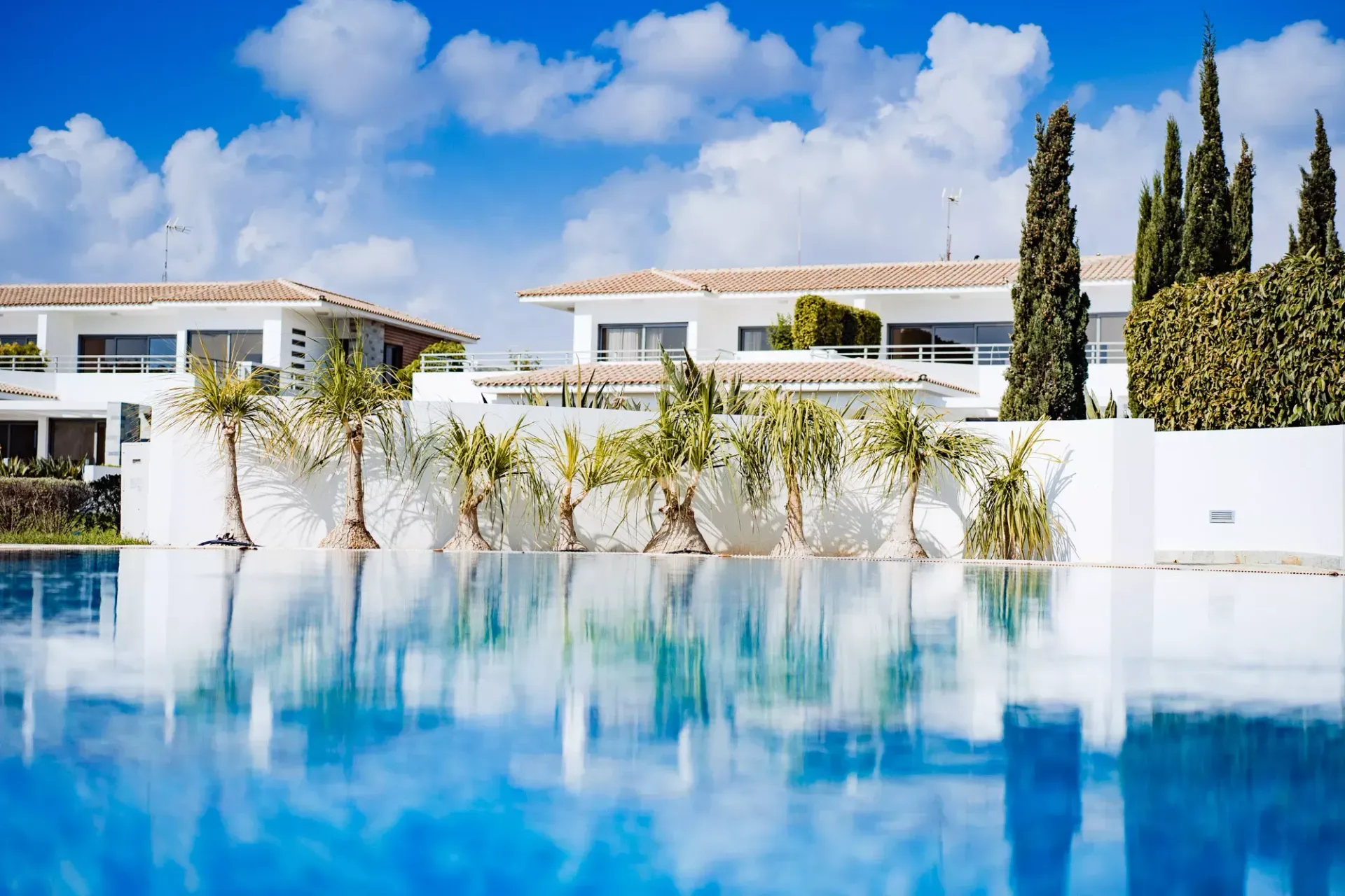 Pool reflecting white buildings and blue sky with clouds; palm trees.